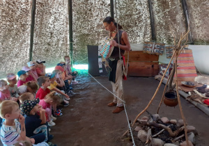 Dzieci siedzą na ławeczkach, a indianin pokazuje i opowiada o wyposażeniu Tipi