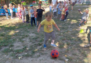 Dzieci stoją i wykonuja kolejno strzał do bramki