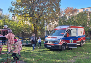 Ambulans stoi w ogrodzie przedszkolnym dzieci oglądają go w środku, włączają sygnały dźwiękowe i świetlne