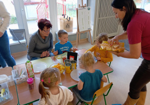 Dzieci siedzą przy stoliku, Babcia rozdaje upieczone gofry