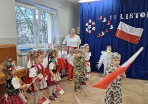 DZieci śpiewaja piosenkę o fladze, chłopiec w stroju żołnierza chodzi po scenie z dużą flagą Polski