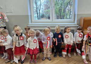 Dzieci najmłodsze na stojąco słuchają piosenki śpiewanej przez starszych kolegów i koleżanki. W dłoni trzymają flagi Polski i nimi machają