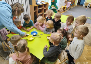 Nauczycielka rozgniata ząbki czosnku w prasce, dzieci siedzą przy stoliku i obserwują działanie