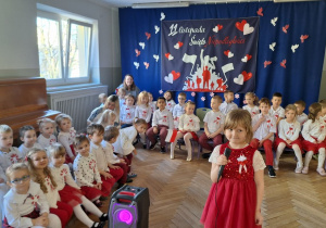 Dzieci ubrane na biało-czerwono siedzą na ławeczkach, dziewczynka rozpoczyna uroczystość