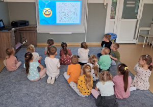 Dzieci siedzą na dywanie i oglądają film edukacyjny związany z dniem kropki