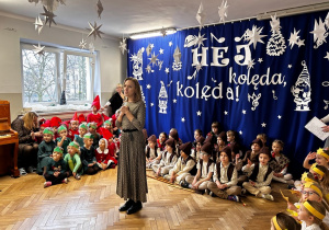 Pani Dyrektor wita zebranych gości, za nią na scenie siedzą w grupach dzieci