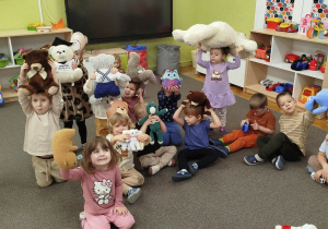 Dzieci siedzą na dywanie trzymając w górze swoje maskotki