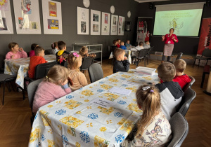 Dzieci siedzą przy stolikach i słuchają prowadzącej, która opowiada o Żołnierzach