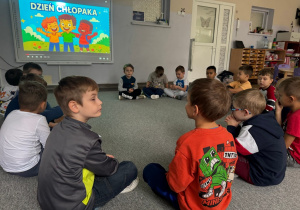 Dzieci siedzą w kole i oglądają prezentację na temat Dnia Chłopaka